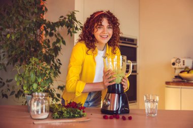 In the kitchen. Curly-haired girl looking involved while making smoothie in the kitchen