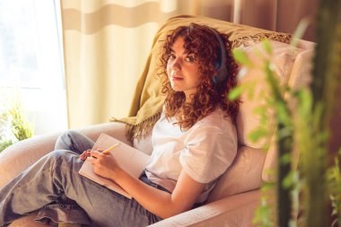 Diary. Curly-haired young girl in headphones sitting in the armchair and making notes in a diary