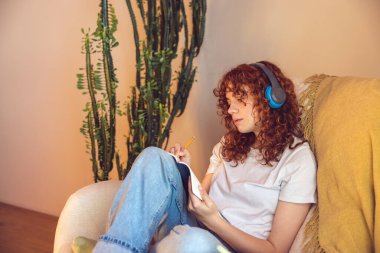 Diary. Curly-haired young girl in headphones sitting in the armchair and making notes in a diary