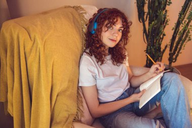 Diary. Curly-haired young girl in headphones sitting in the armchair and making notes in a diary