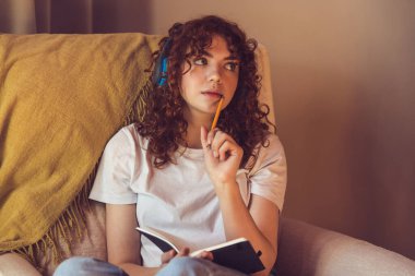 Making notes. Cute girl in headphones sitting in the armchair with diary in hands
