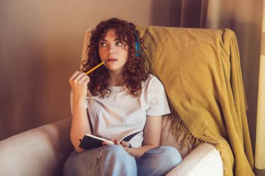 Making notes. Cute girl in headphones sitting in the armchair with diary in hands
