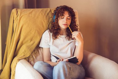 Making notes. Cute girl in headphones sitting in the armchair with diary in hands