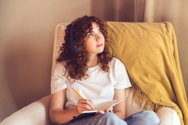 Diary. Curly-haired young girl in headphones sitting in the armchair and making notes in a diary