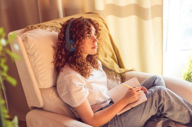 Diary. Curly-haired young girl in headphones sitting in the armchair and making notes in a diary