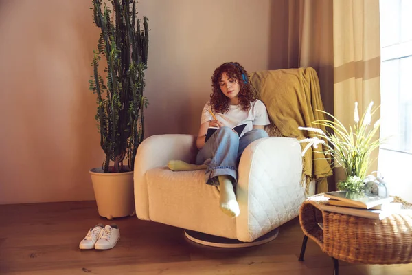 Making notes. Cute girl in headphones sitting in the armchair with diary in hands