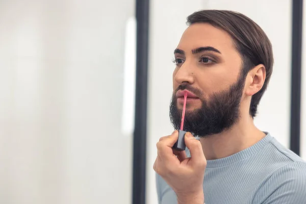 Portrait of a male person coloring lips with a pink lip gloss in the room