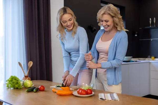 Smiling mature lady seasoning the salad with pepper while a young woman cutting fresh cucumbers