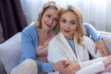 Mutlu yetişkin kızını kucaklarken koltukta oturmuş gülümseyen olgun sarışın kadın.