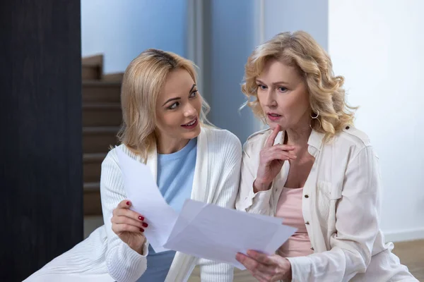Smiling young blonde woman and a serious mature lady sitting on the floor with documents in their hands