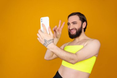 Gülümseyen, sakallı, transseksüel, sarı bluz giyen, selfie çeken, turuncu arka planda V işaretini gösteren bir jest yapan biri.