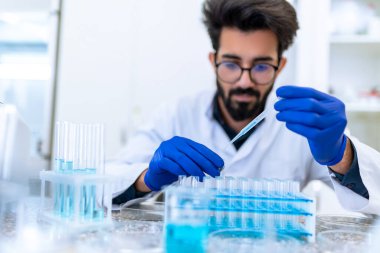 Gözlüklü ve cerrahi eldivenli sakallı bilim adamı laboratuarda cam, bilim ya da kimya araştırma ve geliştirme ile kimyasal numuneyi test ediyor..