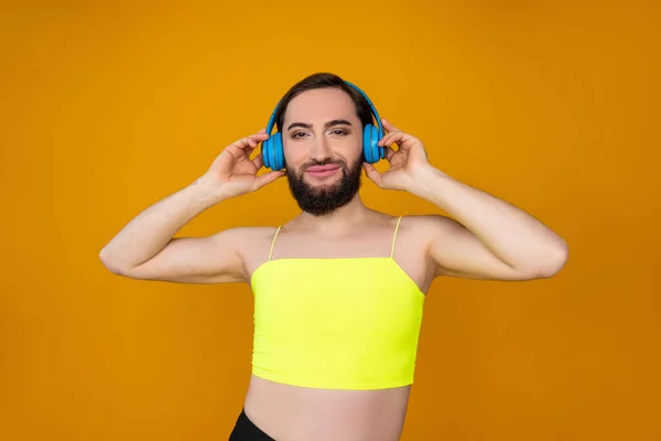 自信に満ちた楽しいですトランスジェンダー人とともに髭を身に着けています黄色トップリスニング音楽を保持手にヘッドフォン上の孤立したポーズオレンジ色の背景 — ストック写真