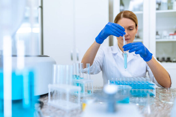 Успешная блондинка-исследователь, работающая с медицинскими образцами, держащая пробирку с голубой жидкостью, заставляя исследовать микробиологию в лаборатории.
