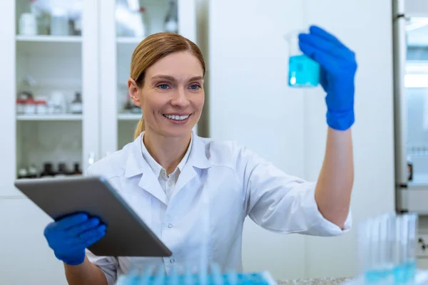 白种人女实验室科学家在她的剪贴板上做一些样品和笔记 拿着蓝色液体瓶 做化学实验 — 图库照片