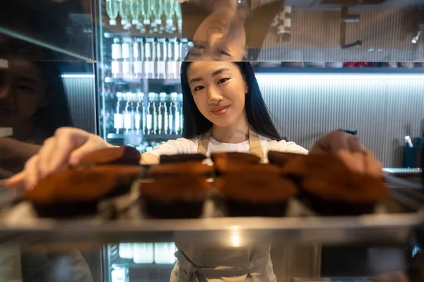 Waist-up portrait of a pleased Asian cafe worker leaning on the glass pastry display case shelf