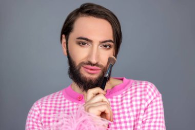 Pembe elbiseli güzel transseksüel insan modeli yüzünü pudralıyor. Gri arka planda izole edilmiş poz veren fotoğraf çekimine hazırlanıyor.