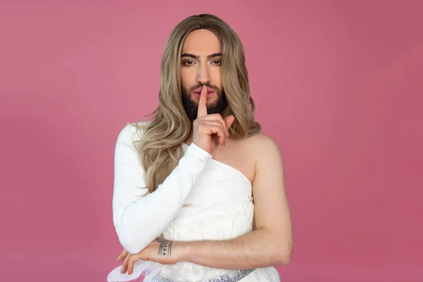 Stock image Mysterious transgender wearing blonde wig and white dress holding hand near lips, keeps secret posing isolated over pink background