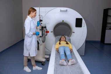 Hastanede MRI taraması sırasında uzun tıp masasında yatan küçük bir hasta, bir klinikte çocuğun tomografisini çeken bir doktor..