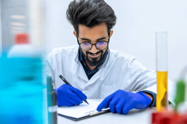 研究室の試験管内の液体を調べる科学者 研究室のコートとゴム手袋を身に着けている彼の職場に座っている — ストック写真