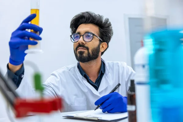 研究室の試験管内の液体を調べる科学者 研究室のコートとゴム手袋を身に着けている彼の職場に座っている — ストック写真