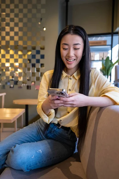 一个快乐的亚洲女人坐在咖啡店的沙发上 用她的手机 — 图库照片