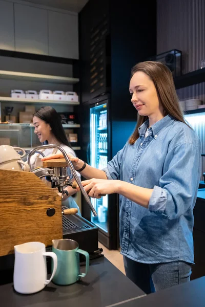 在她微笑的亚洲女同事面前 一位专注的高加索咖啡店工作人员正在操作咖啡机 — 图库照片