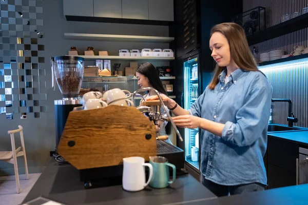 认真专注的白人咖啡师在她快乐的亚洲女同事旁边操作咖啡机 — 图库照片