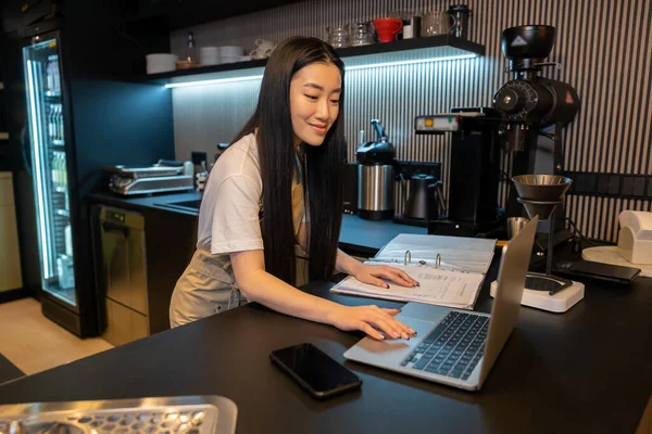专注于咖啡店的年轻员工在手提电脑上工作时触摸账簿 — 图库照片