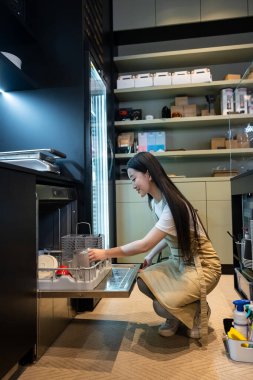 Açık bulaşık makinesinden önce poposunun üzerinde oturan memnun bir kafe çalışanının yan görüntüsü