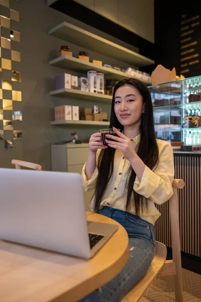 Pleased remote employee with a glass cup of espresso sitting in front of the laptop in a cafe