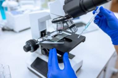 Laboratuvarda mikroskopla çalışan, pipeti mavi sıvıyla tutan, tıbbi eldiven giyen ve genetik araştırmalar yapan tanınamayan bilim adamının görüntüsü..