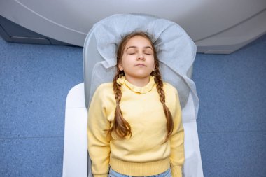 Saçları örgülü küçük kız, tıbbi muayeneden önce gözleri kapalı olarak MRI tarama makinesine giriyor..
