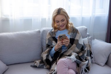 Gülümseyen mutlu kadın rahat koltukta oturmuş elindeki akıllı telefona bakıyordu.