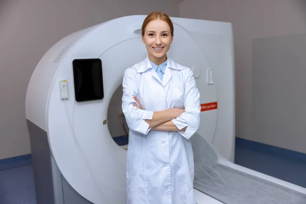 年轻的金发医生站在一家医院的计算机断层扫描机旁边 穿着实验室的外套 对着相机笑着 — 图库照片