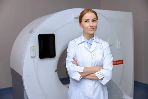 年轻的金发医生站在一家医院的计算机断层扫描机旁边 穿着实验室的外套 对着相机笑着 — 图库照片