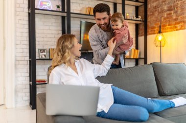 Baba bebeğini taşırken anne bilgisayarını uzaktan kumandalı koltukta kullanıyor koca da karısına bebek için yardım ediyor..