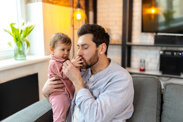 Father with baby, man sitting on sofa with toddler child playing and kissing kids hand with gentle.