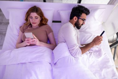 Genç çift karı koca yatakta birbirlerini görmezden gelen telefonlar kullanıyorlar..