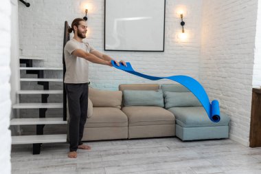 Atletik adam evde egzersiz yapmaya hazırlanırken yoga minderini açıyor..