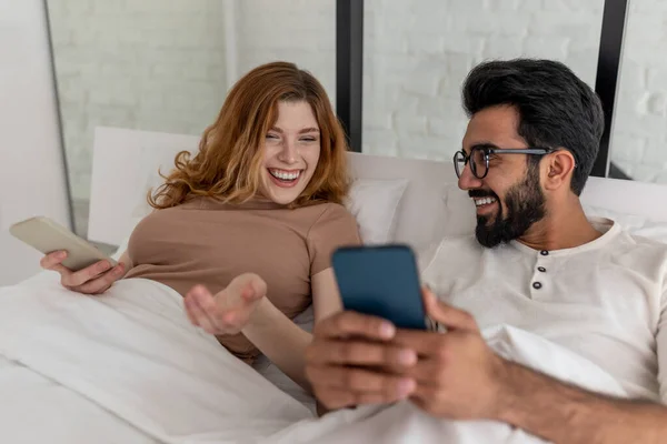 긍정적 남녀가 주말을 침대에서 보내고 핸드폰을 사용하고 즐겁게 웃으며 — 스톡 사진