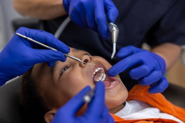 Diş hekimliğinde. Dişçi ve asistanı koyu tenli bir çocukla çalışıyor.