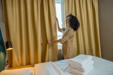 Bir otel odasında. Koyu saçlı kadın otel odasında perdeleri kapatıyor.