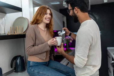 Smiling woman drinking coffee with man while sitting on kitchen counter, husband pouring beverage to her wife. clipart