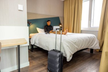 Otel odasında. Genç, siyah saçlı bir adam otel odasında yatıp internette sörf yapıyor.