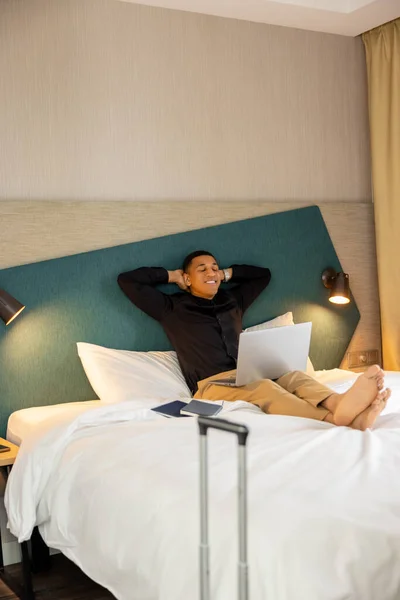 在酒店房间里年轻的黑头发男子躺在床上热腾腾的房间里上网 — 图库照片