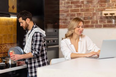 Kadın mutfakta bilgisayar başında çalışırken, erkek kareli gömlekli adam akşam yemeğini hazırlıyor..