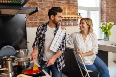 Loving wife and husband preparing dinner, man cooking food in kitchenn and communicating with resting woman. clipart