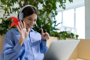 Kulaklıkla gülümseyen genç bir kadının belden yukarı portresi dizüstü bilgisayarın ekranına bakarken baş parmak hareketini gösteriyor.