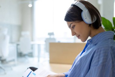 Kablosuz kulaklıklı, belgelere bakan odaklanmış, beyaz bir kadının yan görüntüsü.